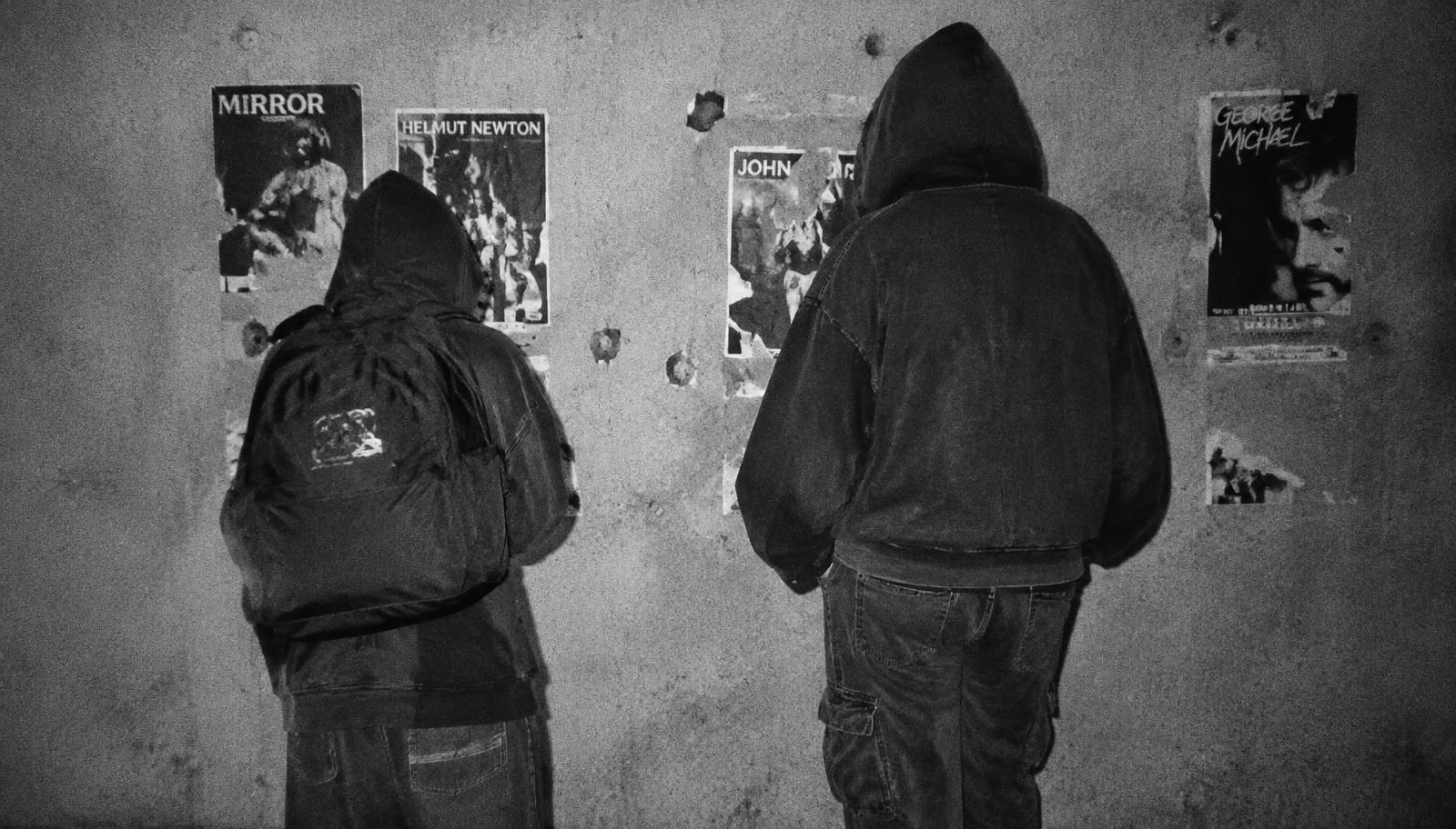 Two figures in oversized streetwear facing a wall of torn flyers